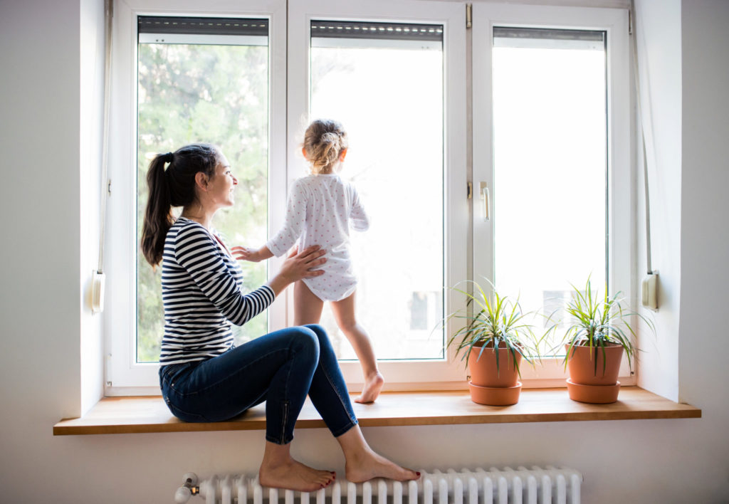 Mother sits on a window sill with child standing on hot water radiator