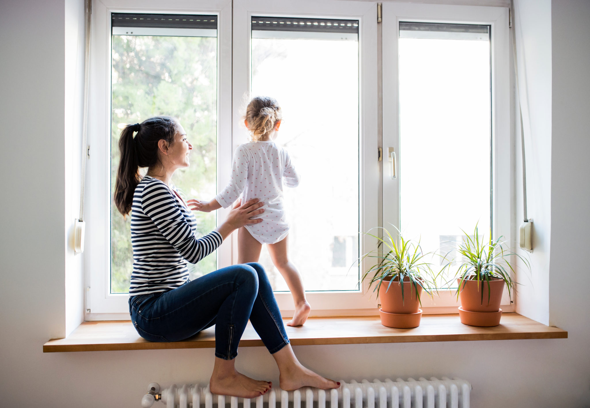 Mother sits on a window sill with child standing on hot water radiator