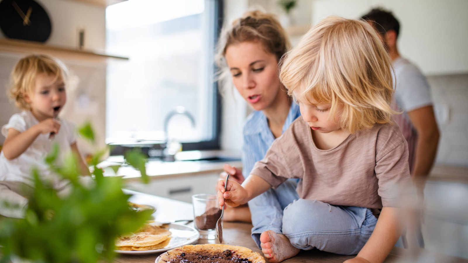 Young family with two small children indoors in kitchen, eating pancakes.